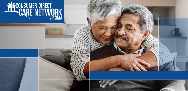 Elderly man sitting on the couch in his living room. Woman standing behind the couch hugging the man.